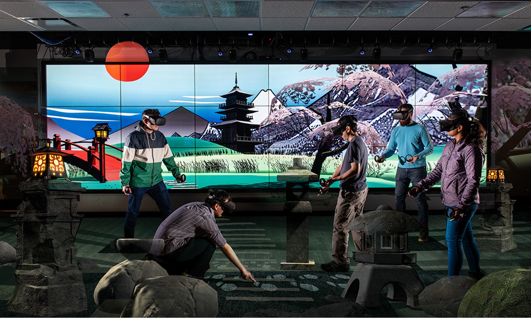 There are 5 people depicted in Virtual Reality head sets. Behind them is a large screen showing a Japanese cherry orchard, a red sun, a bridge and snow-capped mountains.