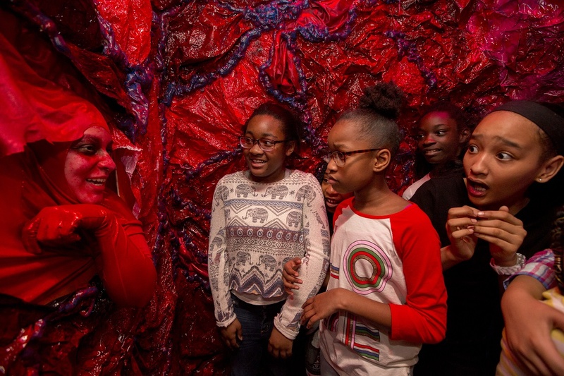  The interior of a human artery with red fleshy walls. A red blood cell appears through the artery wall to address the audience, a group of young kids who are surprised by the blood cells appearance. 