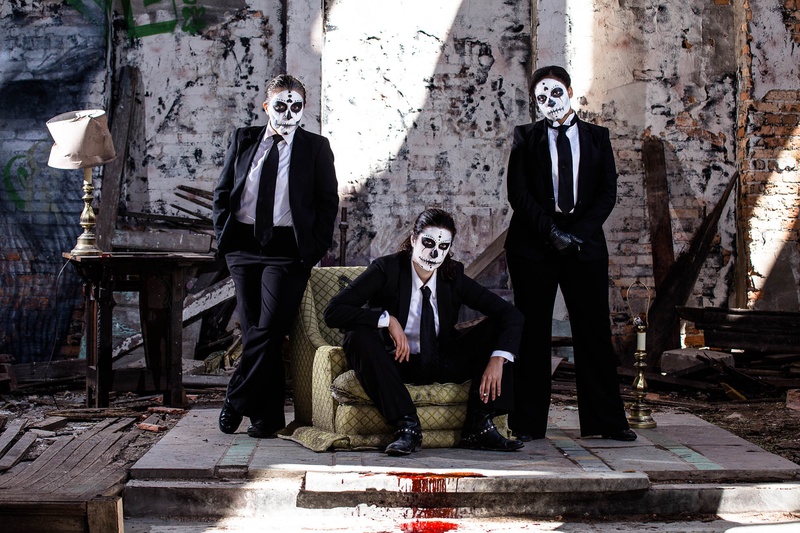  Three intimidating figures, one sitting and two standing by an armchair wearing black suits and day of the dead make up. Blood trails from them and out of the photo. 