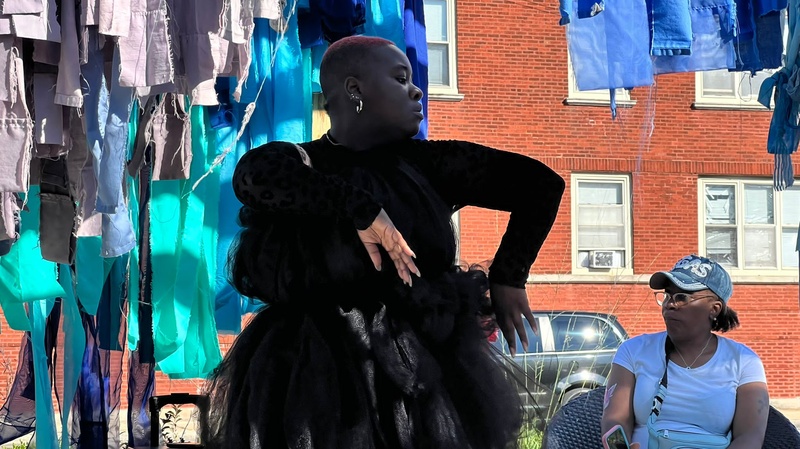  black dark skin woman wearing a long sleeve black body suit under a tulle dress in front of a brick background with blue strips of fabric hanging behind her. 