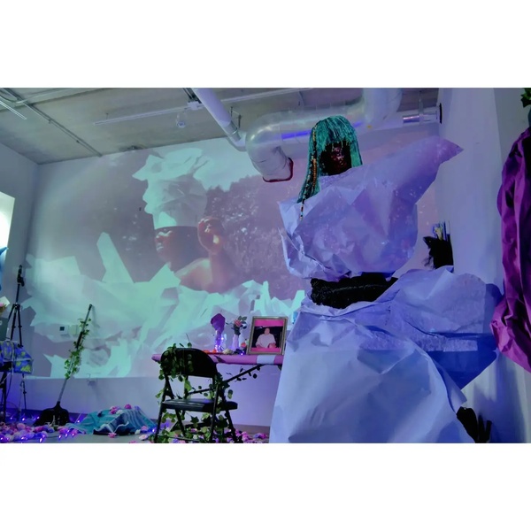  a lavendar paper dressed faceless femme figure in front of a projected image of a black dark skinned woman wearing a white paper dress and head piece with a installation table in from of it 