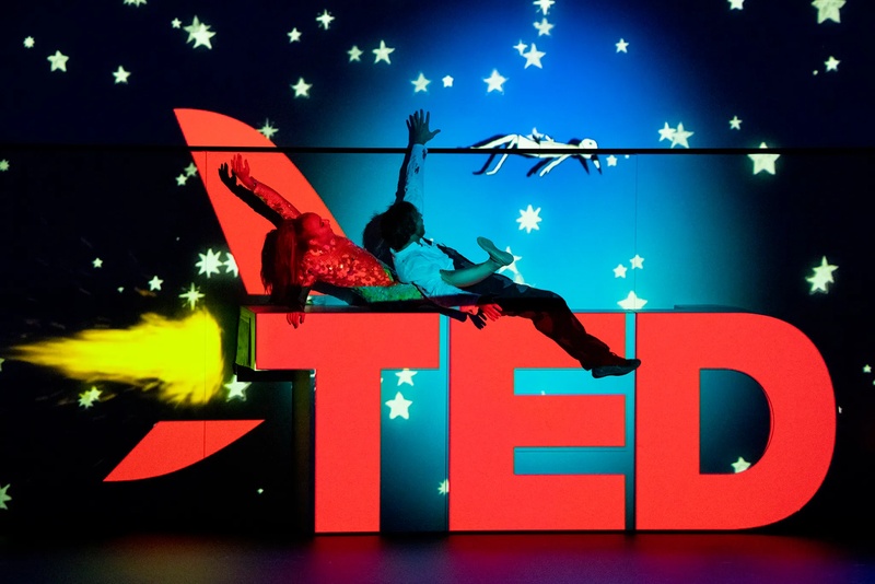 A woman and man riding on top of the red TED logo, which has been turned into a rocket ship and is flying through space 