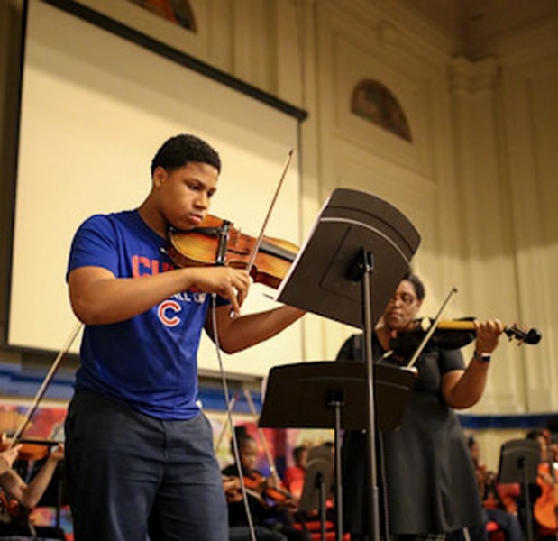  Photo of Ayriole, a brown skinned Black person, standing in the background and playing their violin while in the foreground a student, a brown skinned Black young person with short natural hair and a blue t-shirt with a sports logo, plays their viola. 