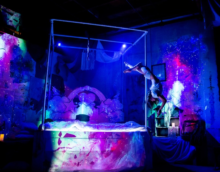  An acrobatic pole dancer spinning and hanging upside down on a pole of a 4-poster bed. Projection mapping paints the image with lilac, pinks and blues. 