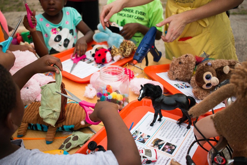  Several children and Marya stand around a tabletop that is covered in toy parts and other items. All are creating toys out of other toys, and most are holding scissors. 
