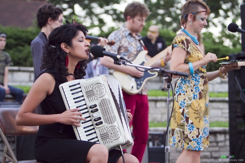  Ami plays a piano accordion outside. She sings soulfully with her fingers on the keys. Behind her are three people playing the xylophone and guitars. Two of them have funky patterned clothing which gives the image some color. 