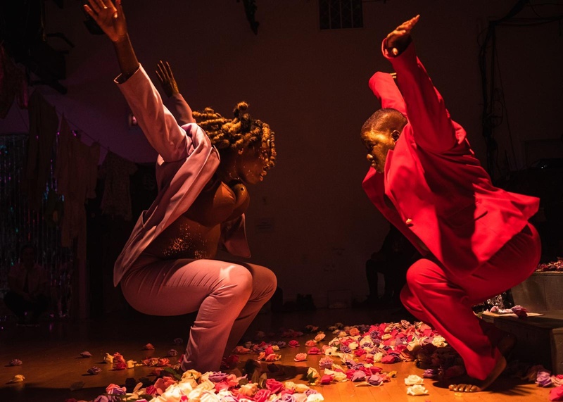  two dark skinned Black women squating down with their arms up in the air facing each other, one in a red suit and one in a lavendar suit, with their faces lightly painted gold, and flowers surrounding their feet 