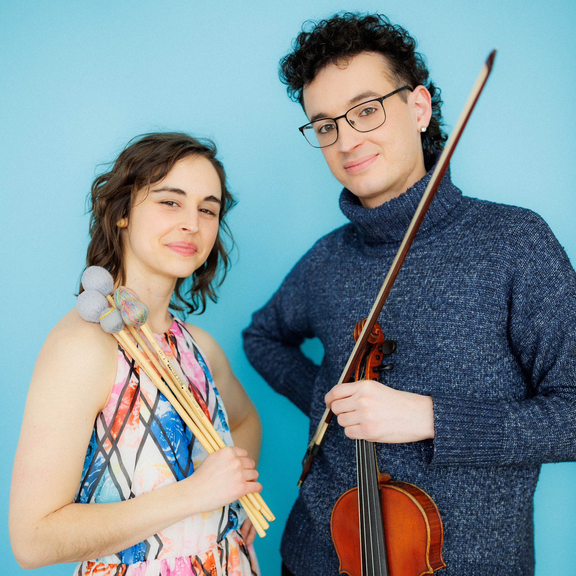  A woman with wavy brown hair and a man with curly dark hair stare at the camera and smile lightly. They are holding mallets and a violin in front of a blue background. 
