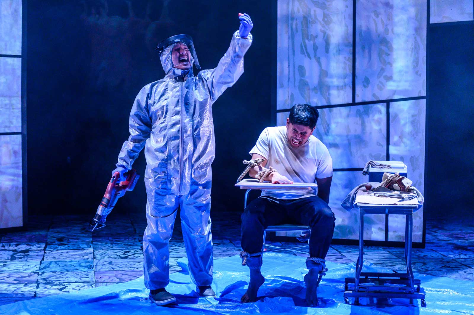 A man in a bio hazard suit holds something in the air while holding a reciprocating saw in the other hand while a man tied to a chair screams.