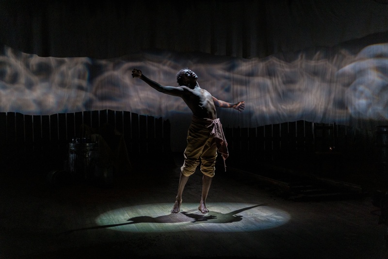 A performer stands in the center of a darkly lit stage. They are shirtless and standing with their arms open.