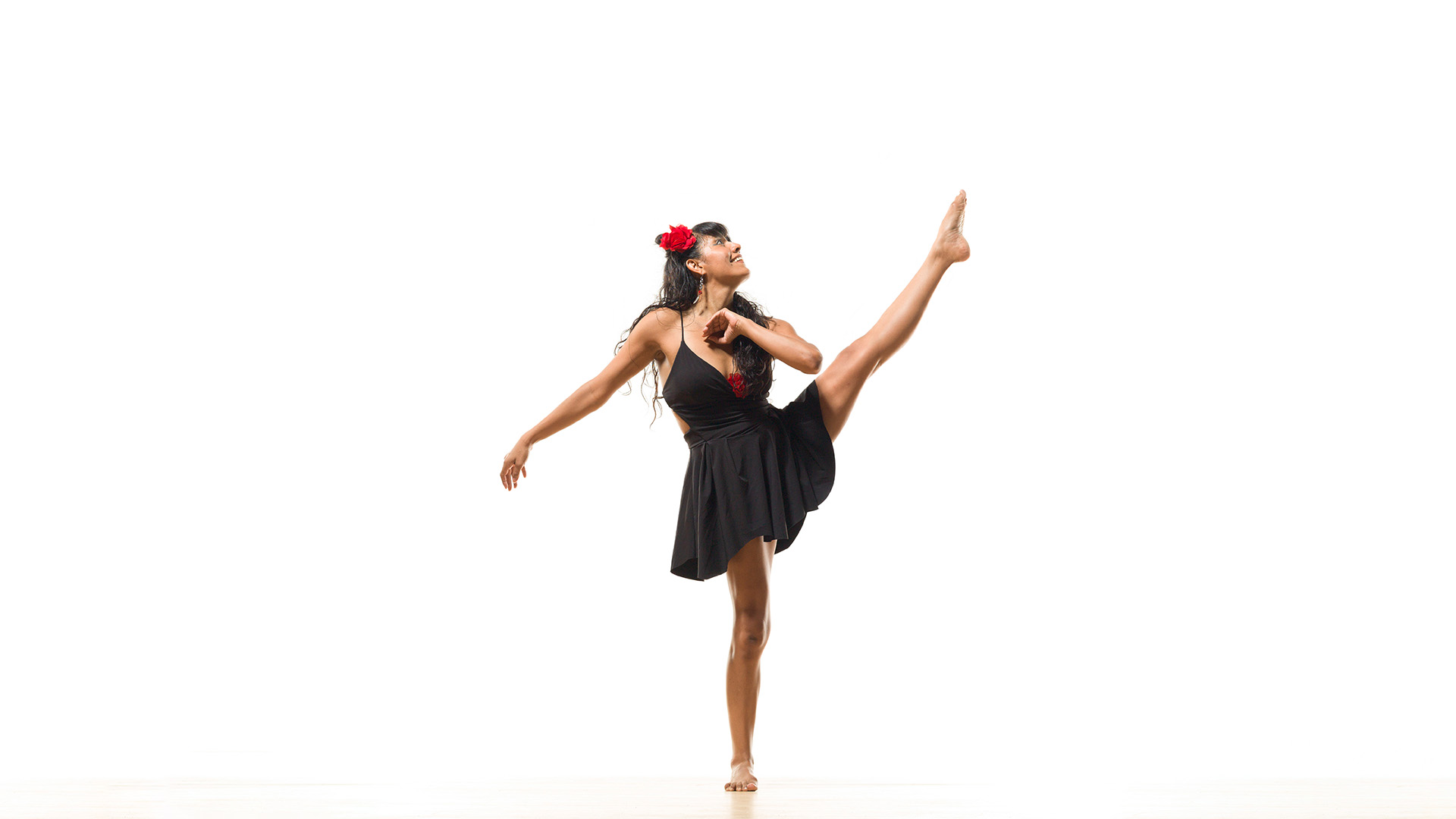 A slender, brown-skinned woman with large brown eyes wears a black dress and a red flower in her hair. She is posing in front of a white background.