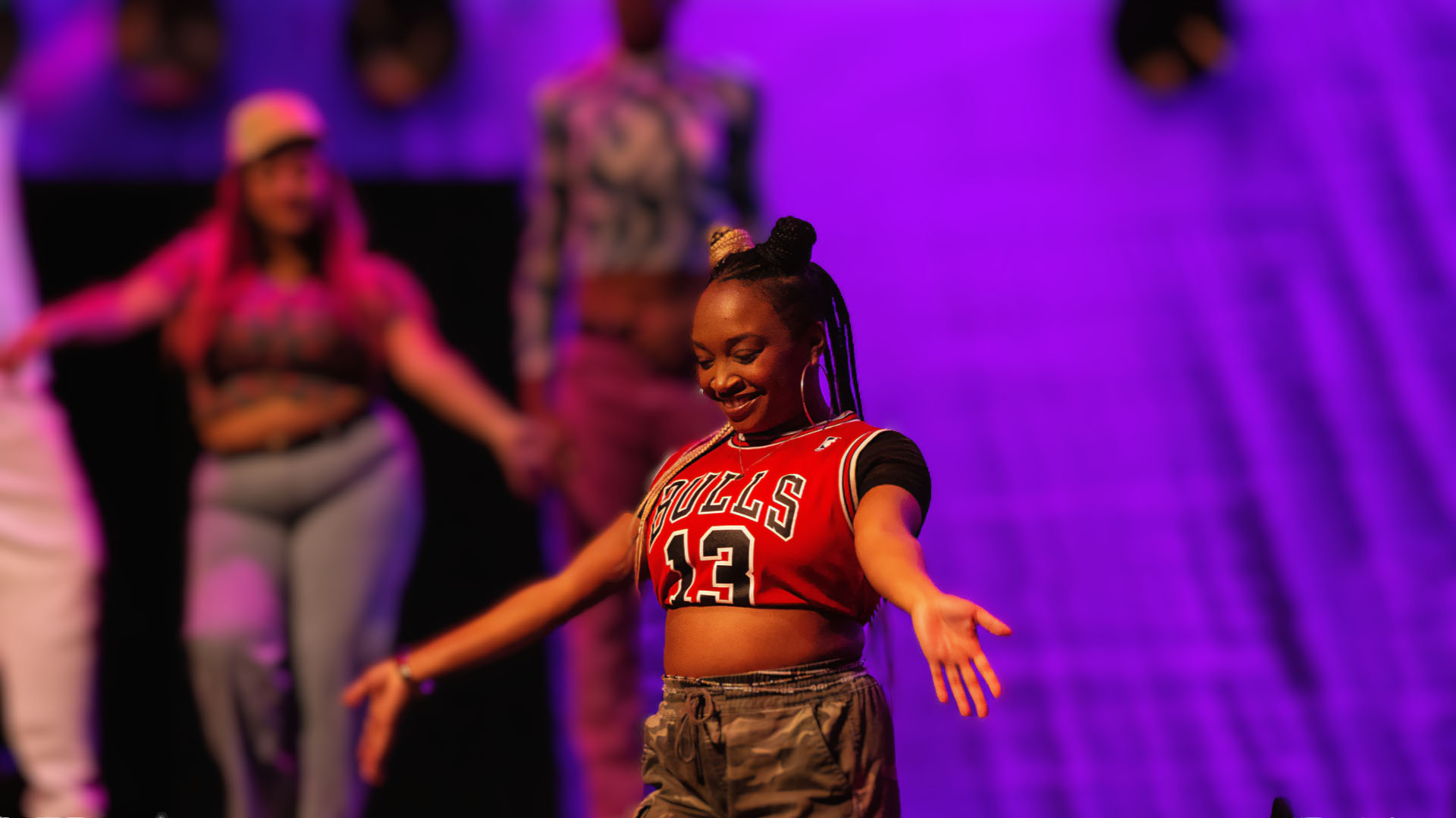 A person dancing with their arms open and smiling in front of a purple background.