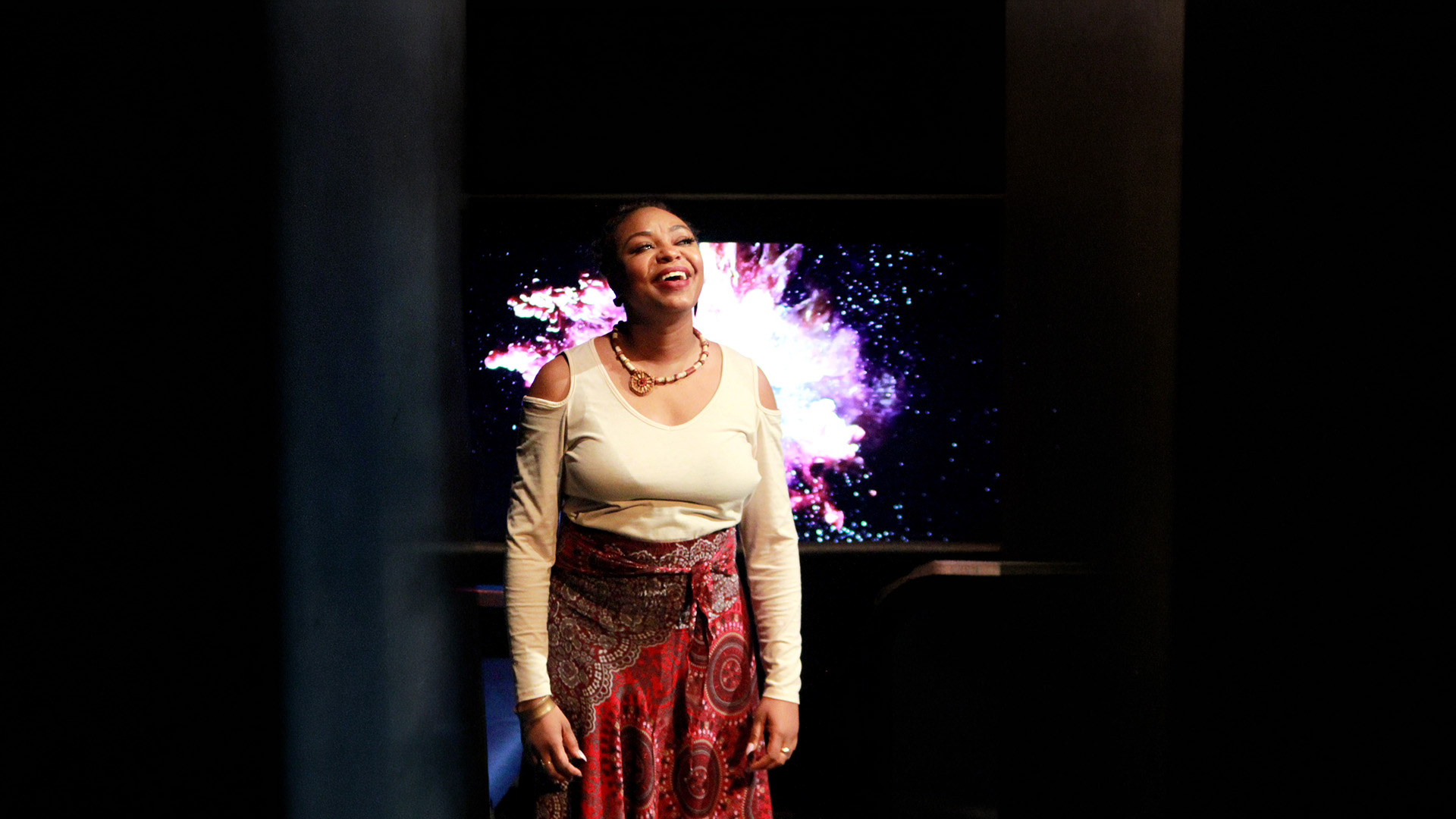 A performer stands on stage smiling. They have short hair and are wearing a white shirt with a beaded necklace.