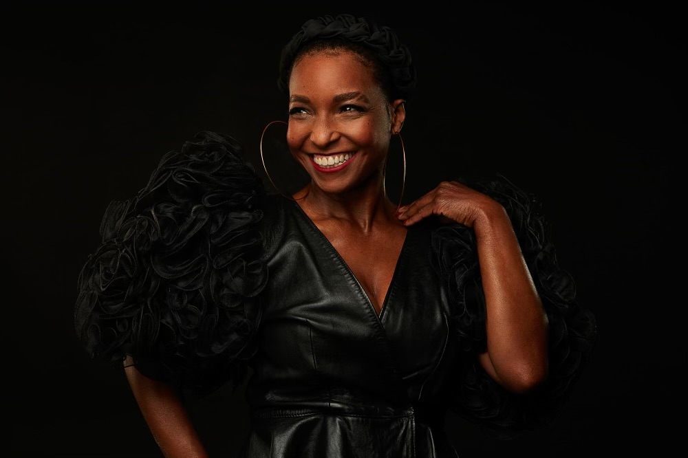 Actor against a black backdrop, also dress in black leather top with large ruffled black sleeves, headband, gold bracelet.
