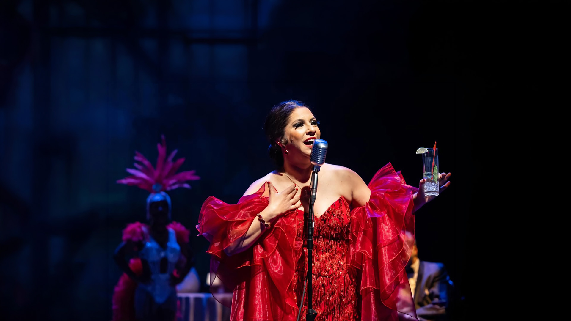 A Latino woman in a red dress performs in front of a microphone.
