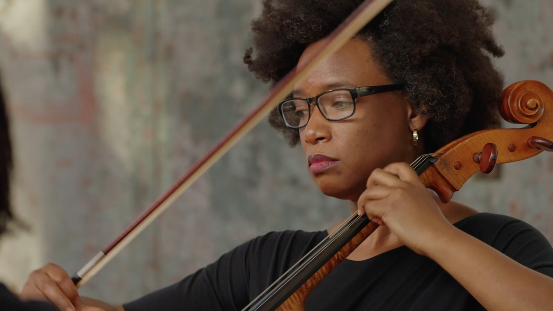 A Black woman with natural hair (Afro) who is smiling. She is dressed in earth-toned colors, wearing an African-inspired necklace and gold hanging half moon earrings. Her lipstick is berry-colored. She has dark brown eyes and is holding a cello.