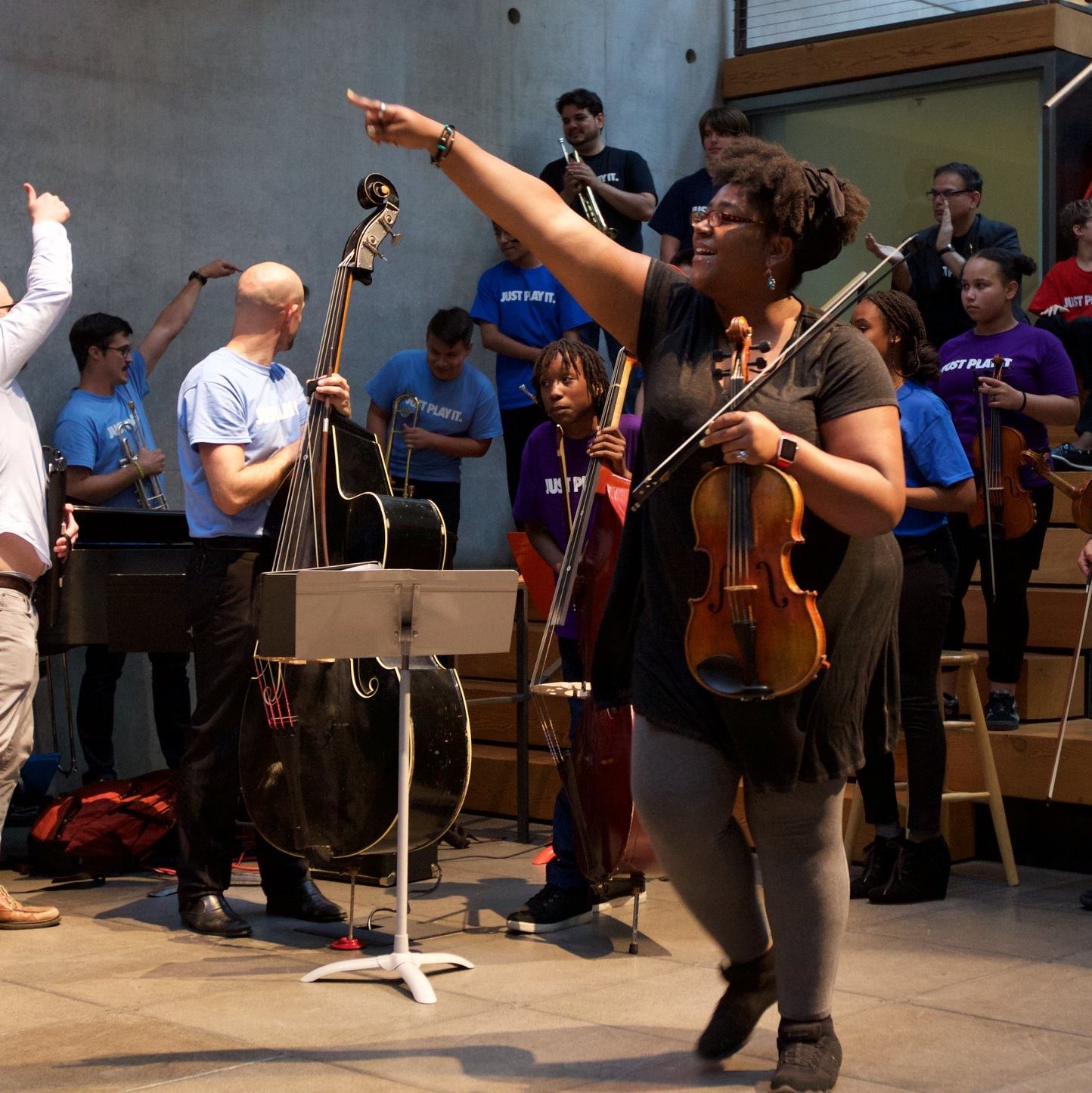 Photo of Ayriole, a brown skinned Black person, wearing a black tunic, grey leggings and black shoes. They stand in a room full of people in bright colored t-shirts, holding string instruments, some on wood risers. Ayriole is pointing and holding a viola.