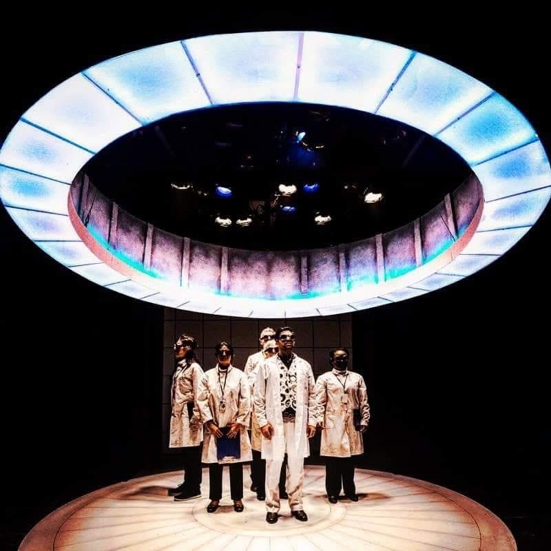 Several people in white lab coats and goggles standing underneath a ring of white-blue light.