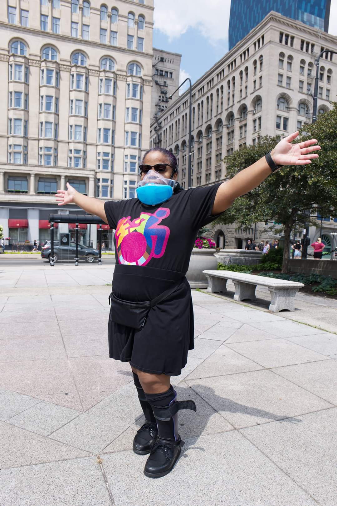 a brown skinned Black woman with spina bifida, standing outdoors in the paved area of a park in downtown Chicago, wearing a PAPR respirator and goggles, a Queer Crip Pride t shirt from Rebirth Garments, a black skirt and socks and black leg braces.