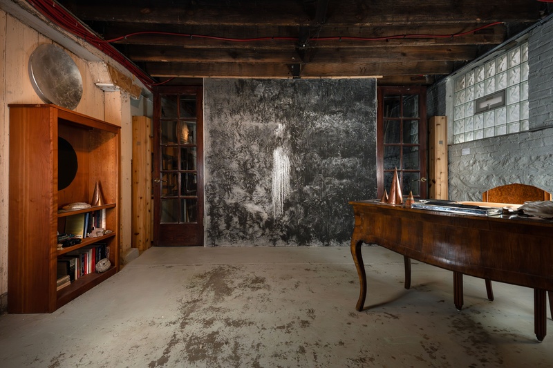  A dimly lit basement features a bookshelf and an antique wooden desk adorned with silver-gilded objects and copper pyramid sculptures. The adjacent wall has a painted mottled grey surface where at the center, water drips into tributaries 