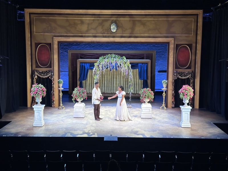  A large gold frame with windows sitting on a marbled floor. Several white planters with flowers sit in a v shape around 2 performers dressed in Victorian style costume. A trellis of hanging flowers is behind them in front of a glowing blue wall. 