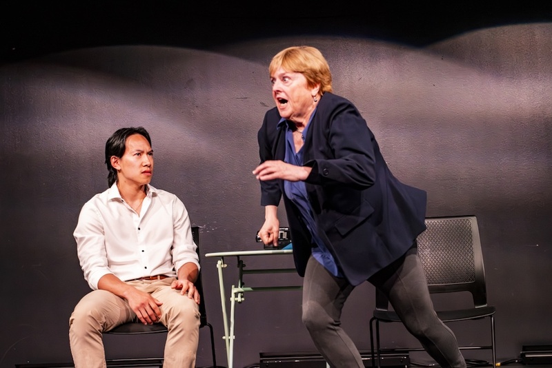  Two performers are on stage. One is sitting in a chair in a white button down shirt while the other is standing up about to run forward. 