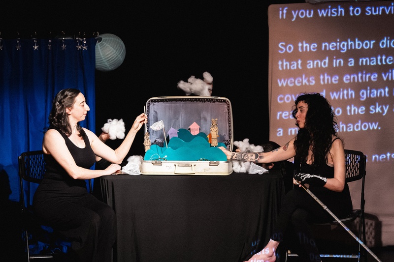  Rachel Singer and Genevieve Ramos sit across from each other at a table.  Between them is a toy theater with hills, houses, clouds and nets emerging from a suitcase. 