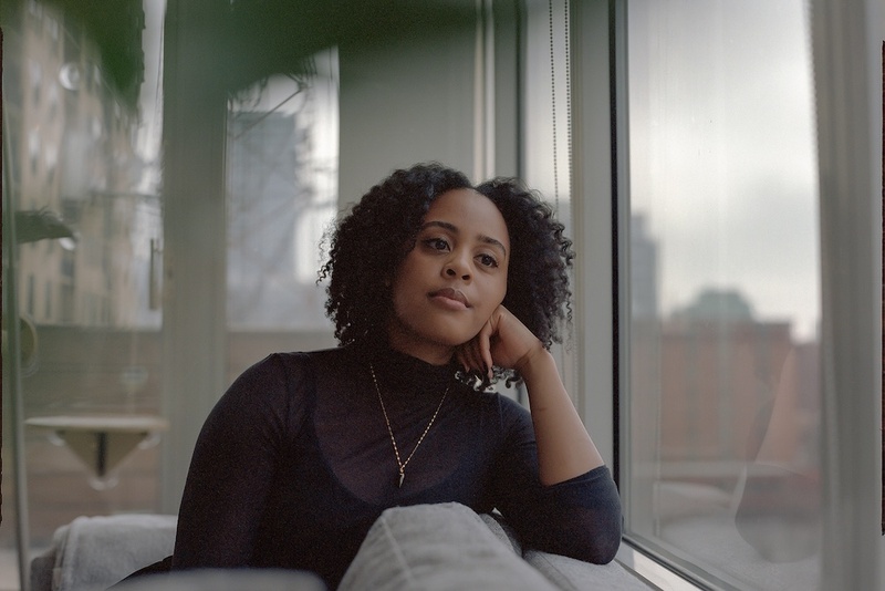  A woman is sitting on a grey couch. She is wearing a black turtleneck and a gold chain necklace. She is resting her head on her hand and has black curly hair. 