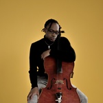 black man with short locs leans on the neck of their cello and looks down to the right