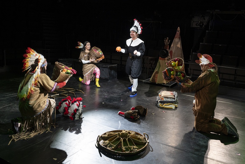  Still from a production for the Thanksgiving play. Three performers are dressed in costume Native American clothing while the fourth is dressed as a pilgrim. The three performers in Native American costumes are holding gourds. 