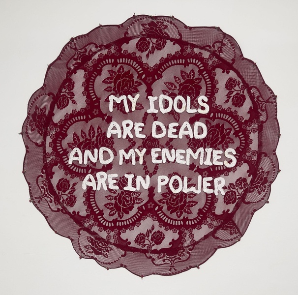  A red lace like textile hanging on the wall. It is round with the words My Idols Are Dead And My Enemies Are In Power stitched in the middle in white thread. 