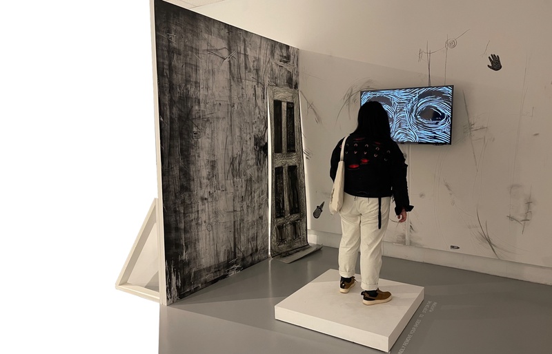  The room is black and white, with only two walls. A TV hangs on the front wall. Charcoal drawings and a black hand-shaped sculpture hang on the white wall. The viewer stands on a white platform to watch the video. 
