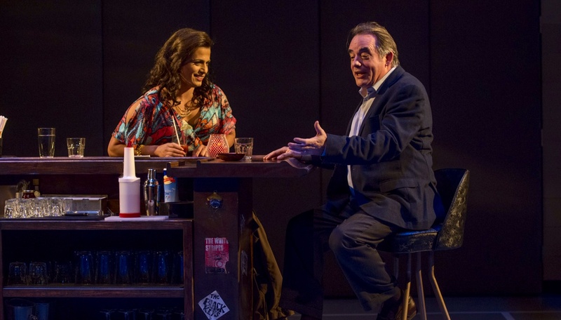 A white man (Tom Irwin) in a dark business suit and beautiful Latina trans woman (Esteban) in a floral print purple, pink and green dress. She has long wavey brown hair. They sit at a bar with drinks and having a lively conversation.