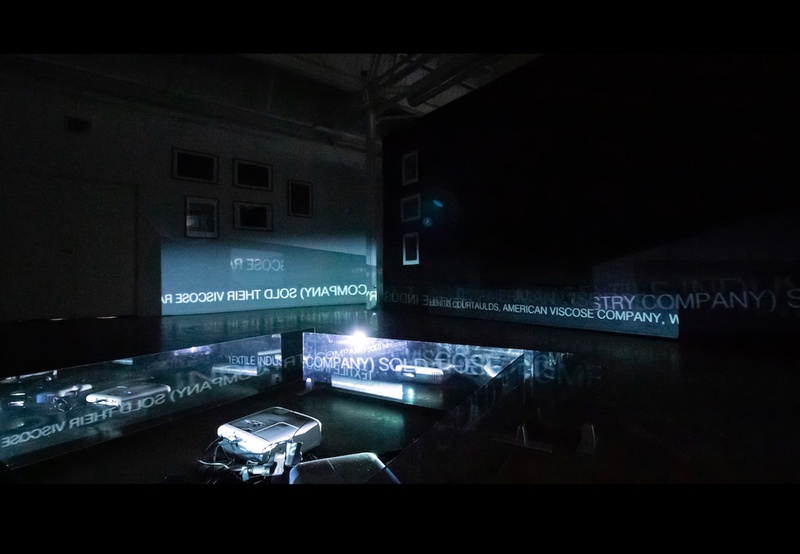  A dark room lit by a projector on the floor. The projector is displaying words on to two way mirror panels. 