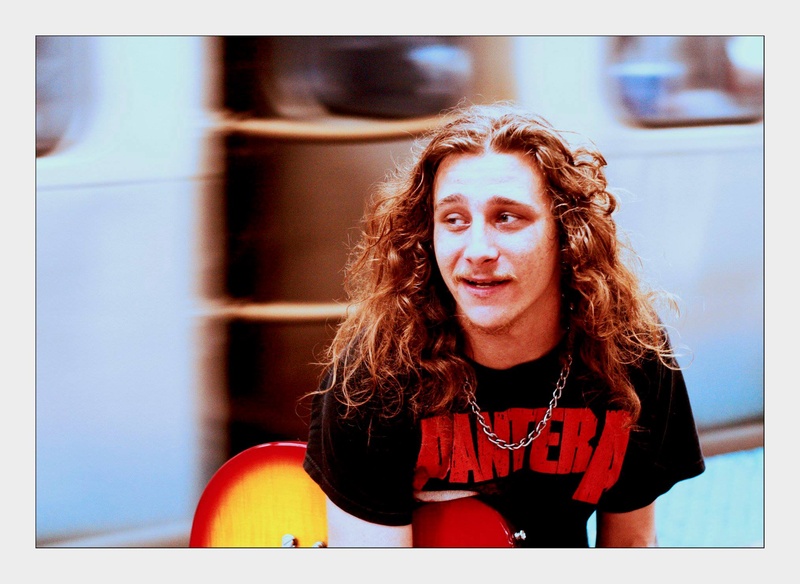  White male guitarist wearing a Pantera shirt sitting in front of a moving L train 