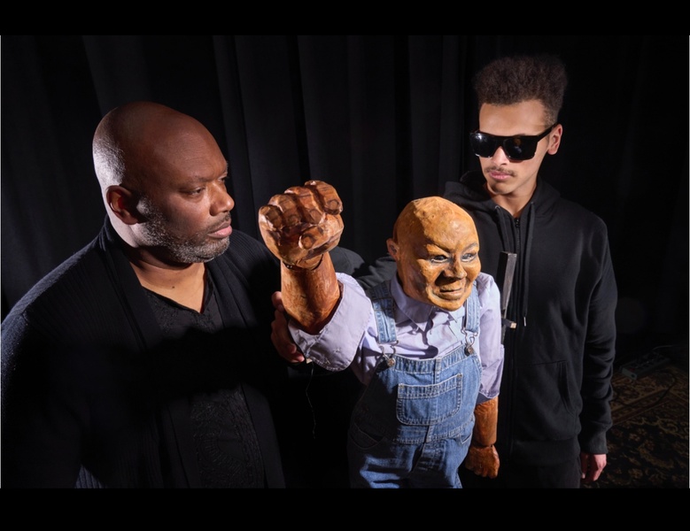  Two performers dressed in black stand holding a puppet carved of wood. The puppet is a man with a blue shirt and overalls on. 