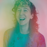 Portrait of a light-skinned person with curly shoulder length hair, smiling at the camera as if caught in the middle of a laugh. Rainbow streaks color the image.