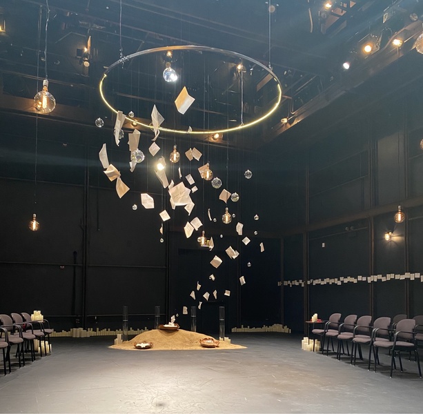  A large space with a hanging gold ring. Several pages float in the air into the ring from a sandbank on a slate floor. 3 wooden bowls with pages sit on the sandback. Pages surround space. Candles, lightbulbs, and pillars of water surround space. 