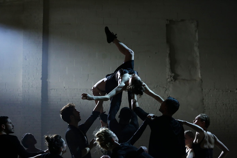  Nine performers stand in a room and lift up one performer in the air. The performer in the air is dressed in black and is twisting around, her back being supported by the others. 