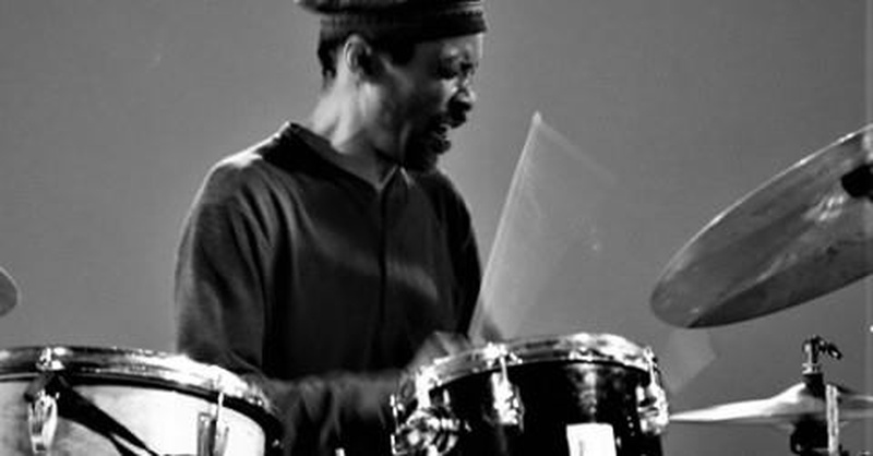  A twinkly brown-eyed African American man with short, curly black hair and a mustache playing drums. 
