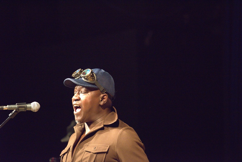  A black man performs on stage behind a microphone. He is wearing a blue hoodie and grey scarf. 