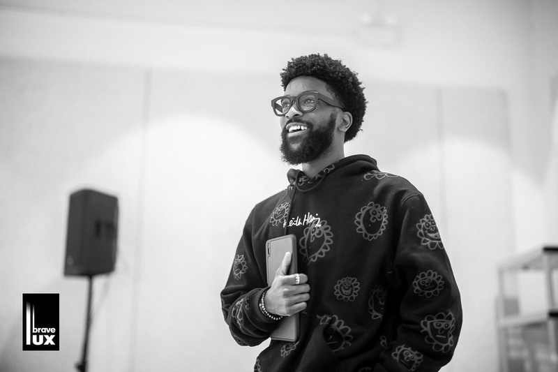  A black and white photo of a black male with short curly hair, a beard and glasses standing in a gallery space. They are holding an iPad and smiling. 