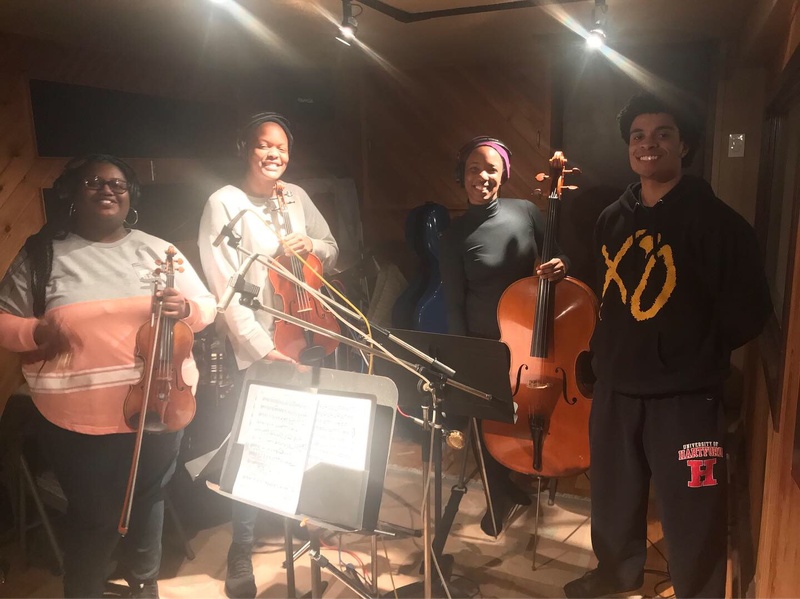  Four musicians stand in a recording studio. One is holding a cello, two are holding a violin. 