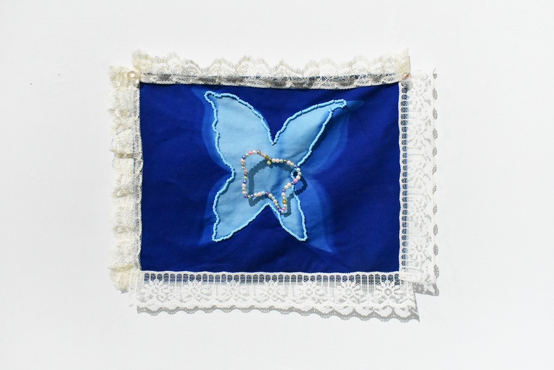  Textile hanging on the all. At the center is a dark blue background with a light blue butterfly, on top of the butterfly is a beaded ring. 