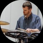 White man with beard and blue prosthetic eyes holds two drumsticks