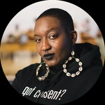 Baty with a short fade, large cowrie shell bamboo earrings, and black lipstick. Her face is slightly tilted as she looks directly into the camera with a half-smile. She is wearing a black hoodie that reads “Got Consent.”