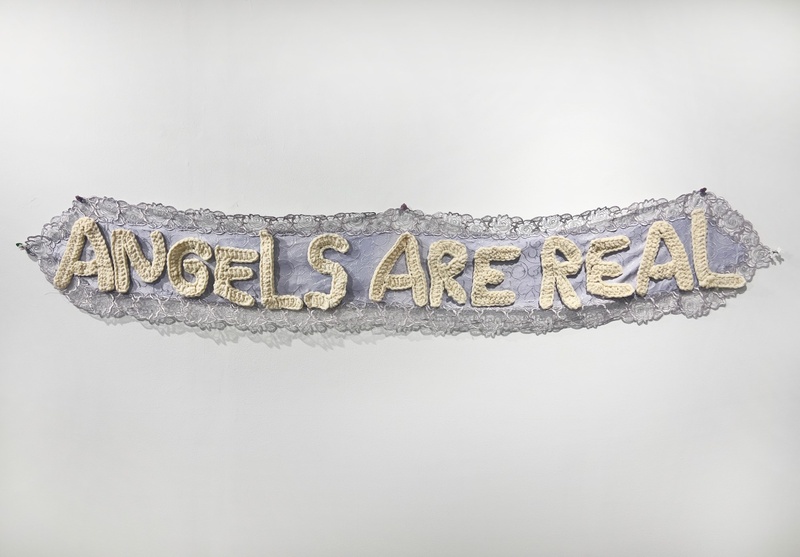  A lace white textile hangs on the wall. On it is stitched in large white crochet the words Angels Are Real. 