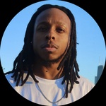 A black man, alethic build with brown locs and brown eyes with Chicago skyline background, blue sky; he use he/him pronouns