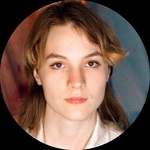 A white, queer transmasculine person sits in front of a brown and blue back drop. They have light brown hair in a shag cut, with brown eyes and fair skin. They are wearing a silky white button up,