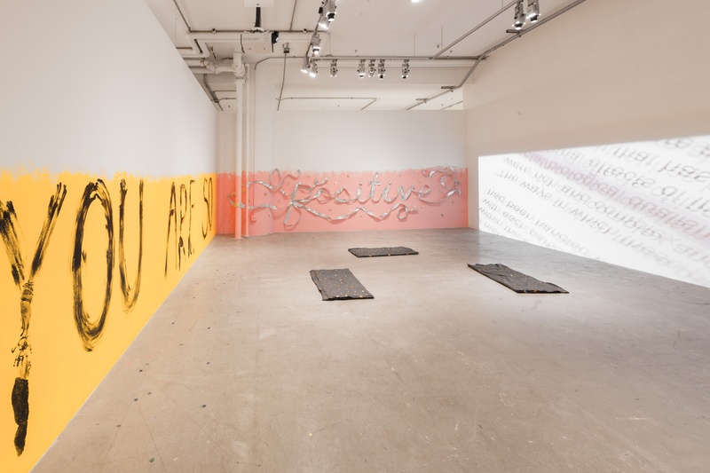 A gallery space. The left wall is yellow halfway up the wall and says the words "You are so". The middle wall is pink half way up and says the word "positive" with ribbon. The third wall shows a a projection with blurry black text. Grey cushions on floor